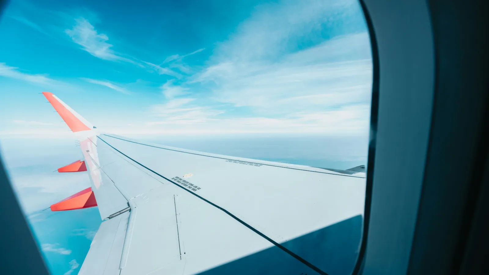 Aircraft wing seen through a passenger window above clouds on a bright international flight