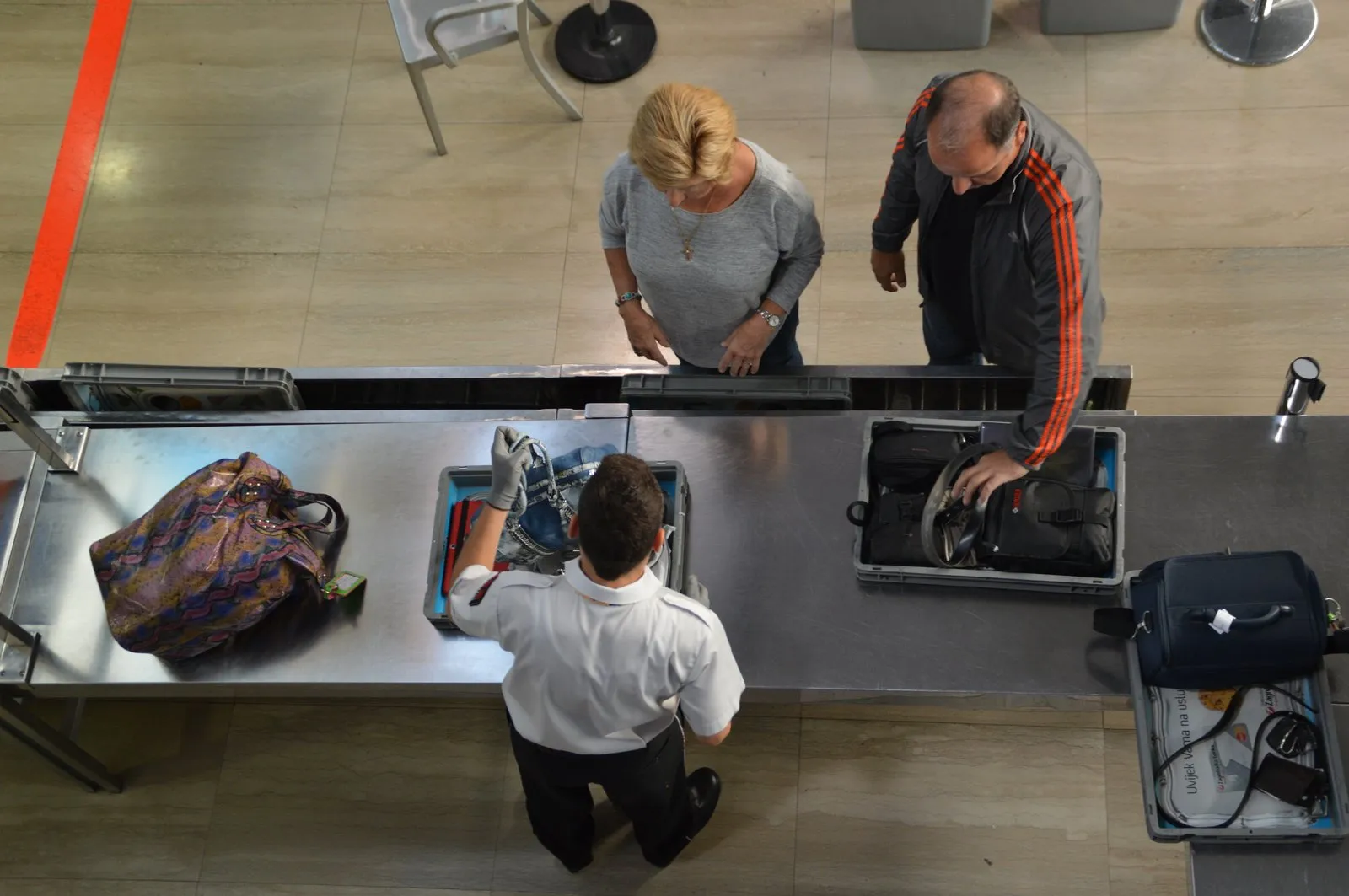 Passengers placing bags in trays while a screening officer checks items at airport security
