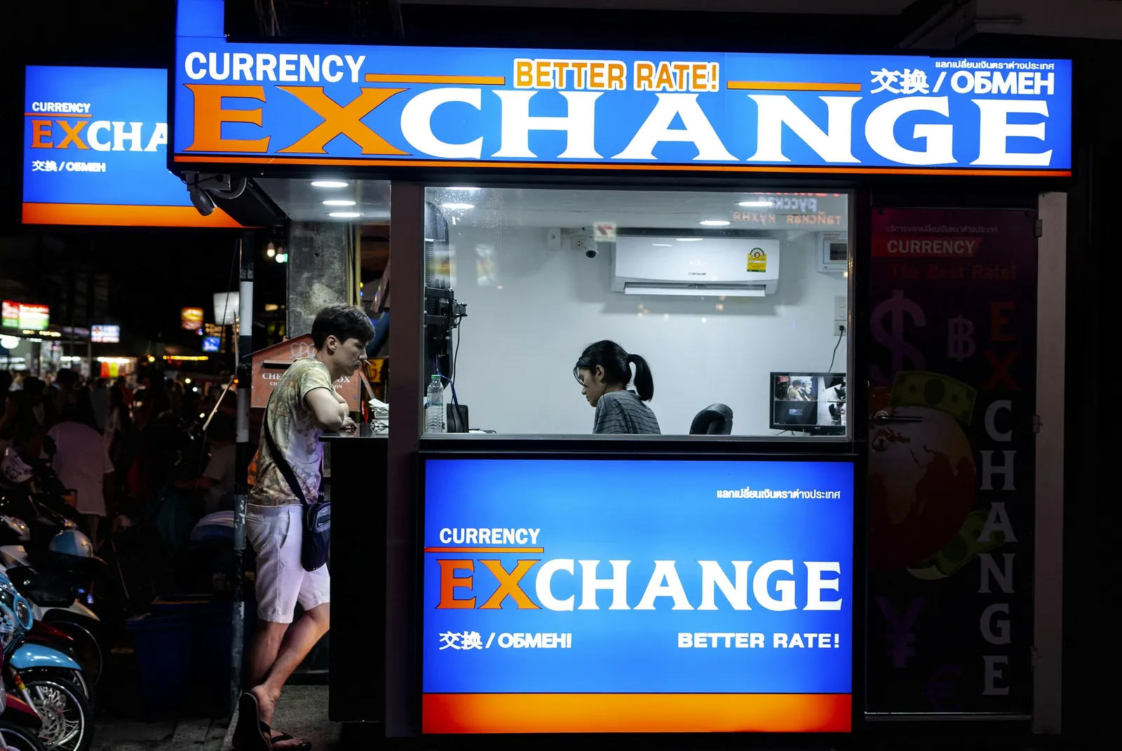 Brightly lit currency exchange booth at night with a customer at the counter window
