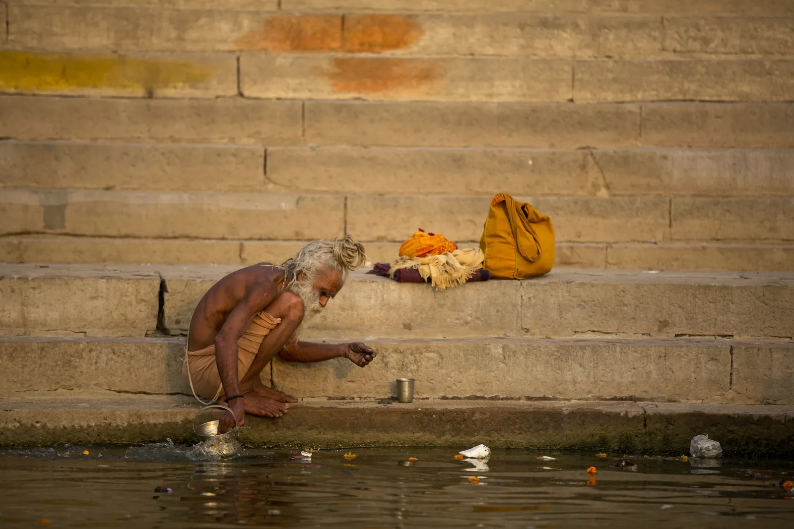 Elderly pilgrim resting on worn stone ghat steps beside the river with his belongings