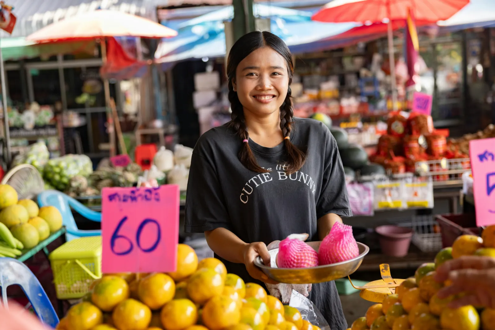 Smiling vendor holding fruit at a Thai fresh market stall piled with oranges and price signs