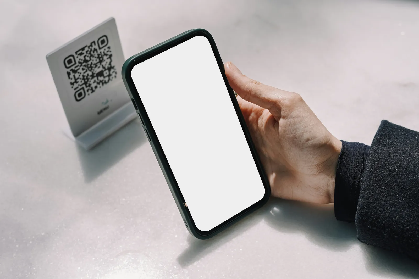 Hand holding a smartphone beside a QR code stand, as when checking an application status online