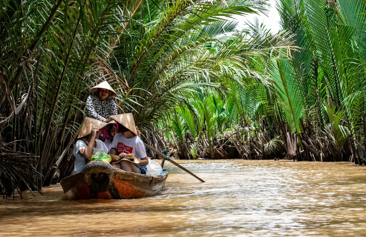 Visitors in conical hats rowing along a palm lined canal in the Mekong Delta