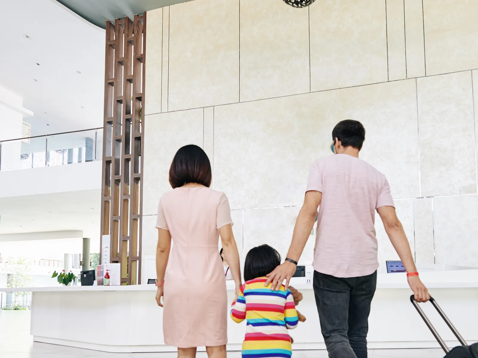 Parents walking hand in hand with a young child and a suitcase through a bright lobby