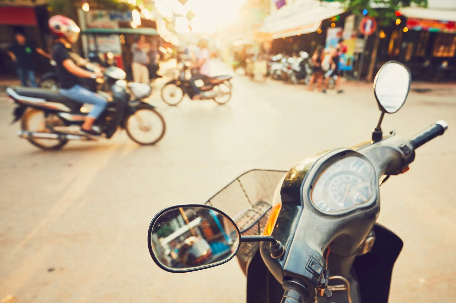 Busy street with motorbike traffic seen past a parked scooter mirror at sunset