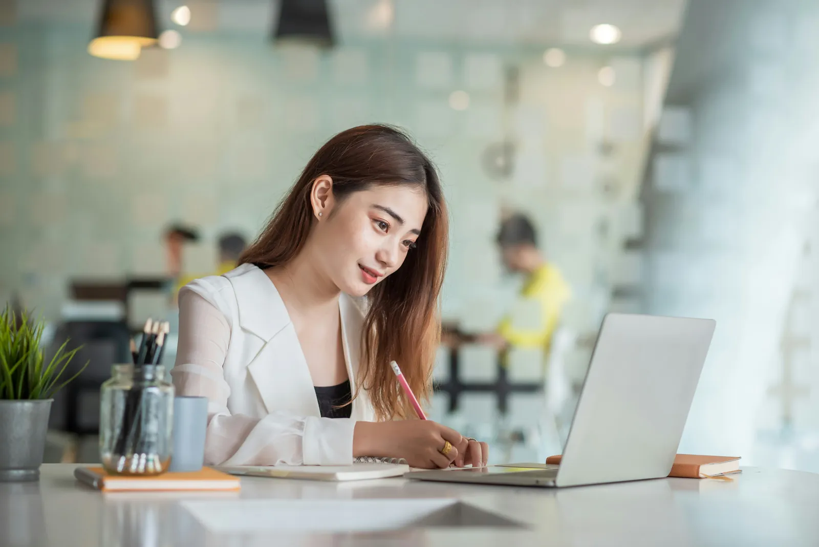Young woman at a cafe table with a laptop, noting details while filling an online form